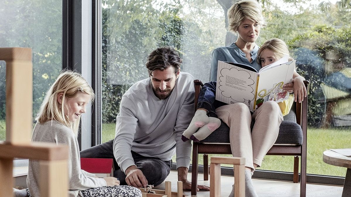 Mann sitzt auf dem Boden und spielt mit einem Mädchen, während eine Frau mit einem weiteren Mädchen auf einem Stuhl sitzt und ein Buch vorliest